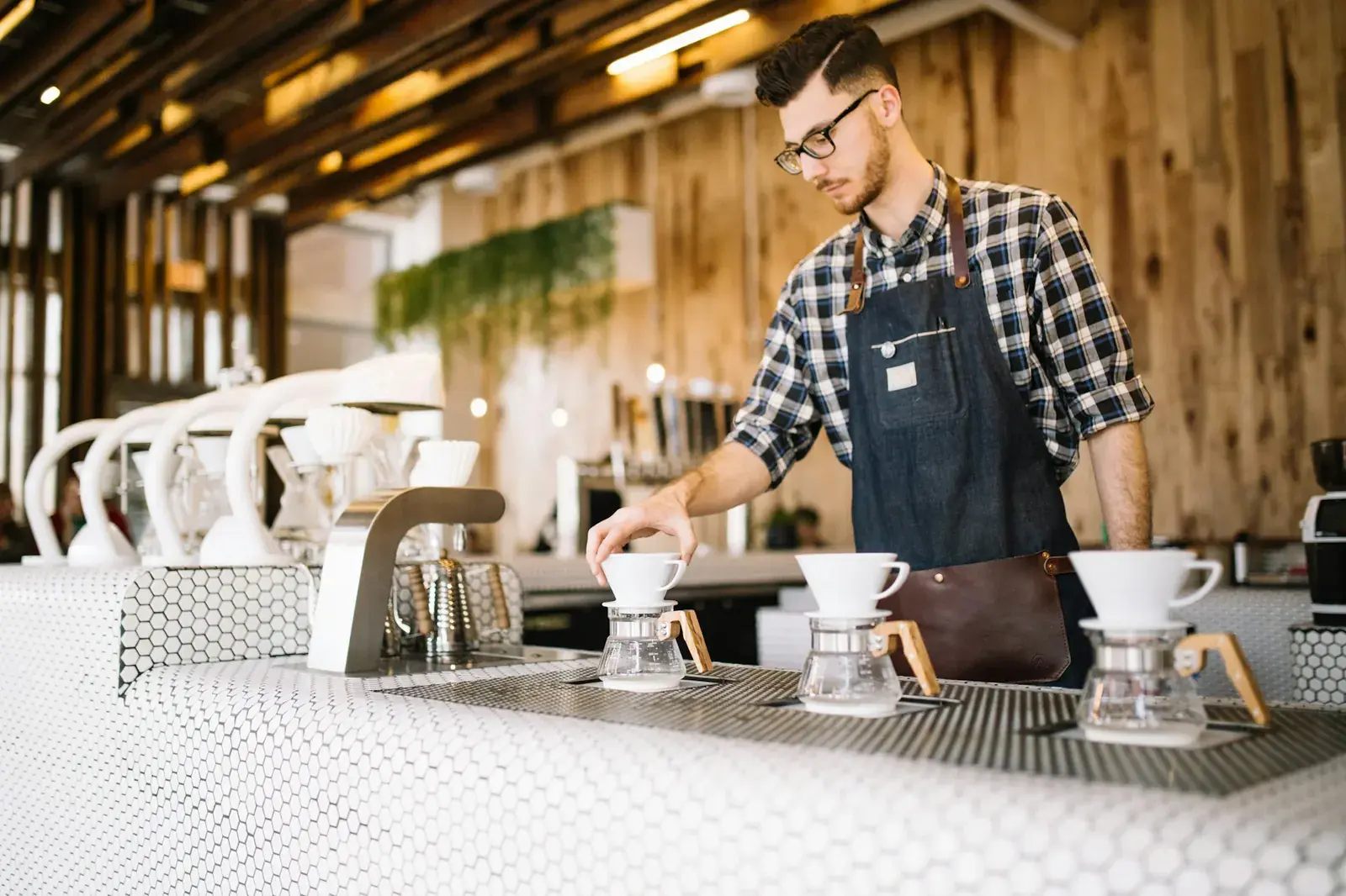 Un giovane barista con occhiali e grembiule prepara il caffè al bancone di un piccolo locale in legno.